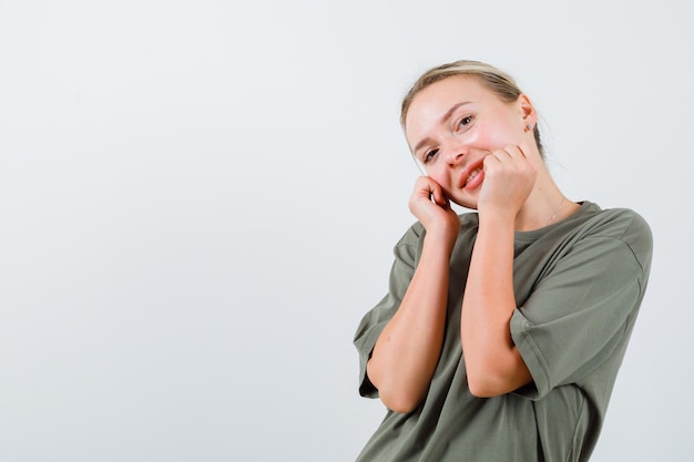 Portrait of lovely blonde in khaki oversize T-shirt puts hand on cheeks and smiles cutely isolated on white background