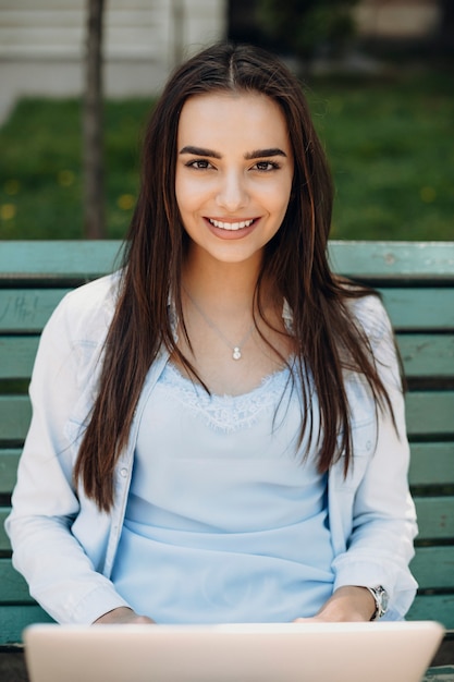 Portrait of a lovely caucasian woman with long dark hair looking directly smiling while sitting on a bench with a laptop on her legs outside.