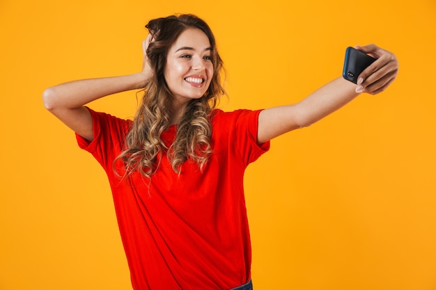 Portrait of a lovely cheerful young woman standing isolated over yellow wall, taking a selfie