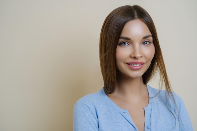 Portrait of lovely dark haired woman with pleasant appearance full lips wears casual blue jumper poses against beige background has calm expression sensual look healthy skin after facial treatment