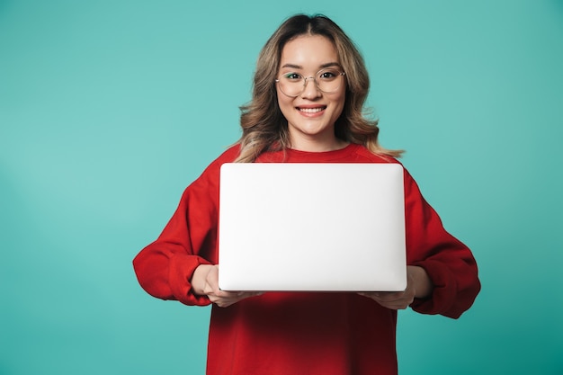 Photo portrait of a lovely excited young asian woman standing isolated over blue wall, using laptop computer