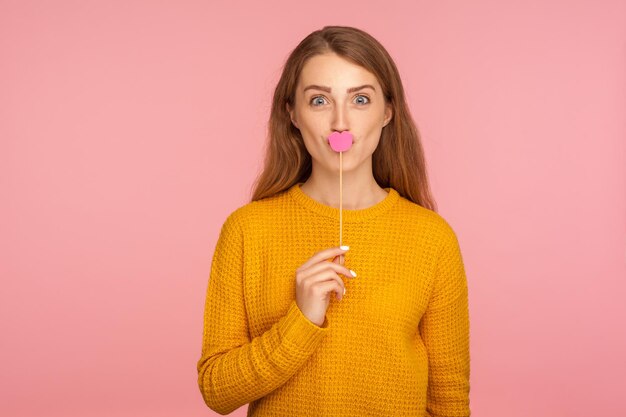 Portrait of lovely funny ginger girl in sweater covering mouth with pink paper lips on stick and looking happily at camera symbol of kiss and love indoor studio shot isolated on pink background