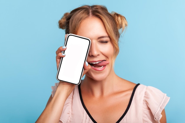 Portrait of lovely positive blonde woman with two funny hair buns in dress standing covering half face with phone and closing eyes, showing tongue. Indoor studio shot isolated on blue background.