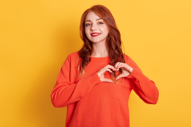 Portrait of lovely red haired female model makes heart gesture, demonstrates love sign, has happy expression, wearing orange sweater, posing against yellow background.