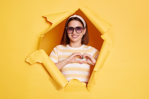 Portrait of lovely romantic woman wearing striped shirt and hair band breaks through yellow paper background showing heart shape with fingers expressing love and gentle looking at camera