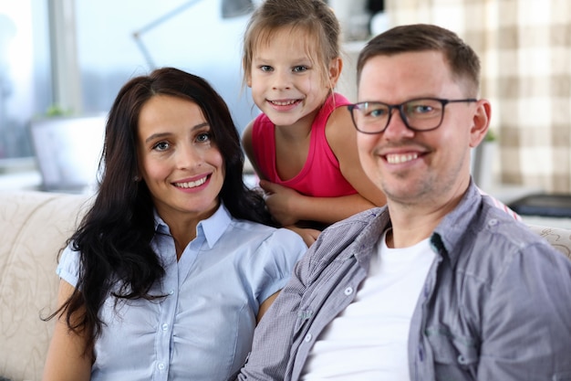 Portrait of lovely smiling family spending time together. Happy mother, father and little child sitting on sofa and looking with gladness. Childhood and parenthood concept