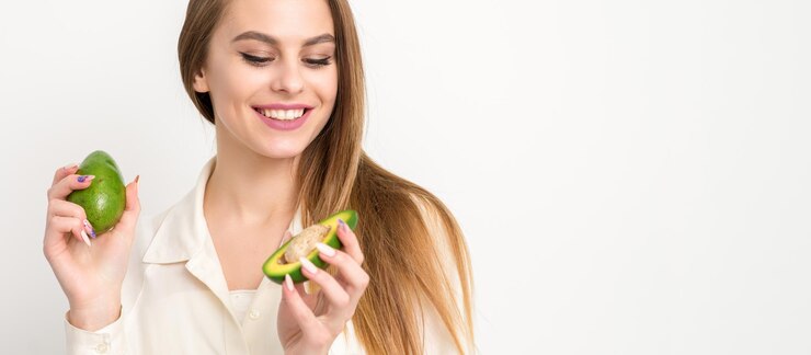 Portrait of a lovely smiling young brunette caucasian woman wearing the white shirt with long hair holding and showing avocado standing isolated over white background