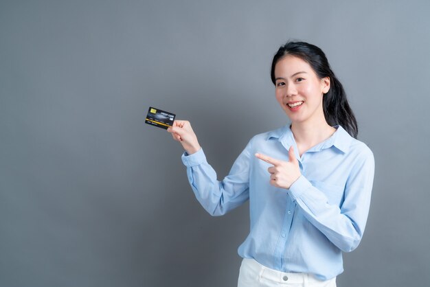 Portrait of a lovely young asian woman in blue shirt showing credit card with copy space on grey wall