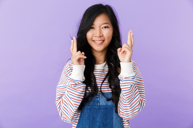 Portrait of a lovely young asian woman isolated over violet space, holding fingers crossed for good luck