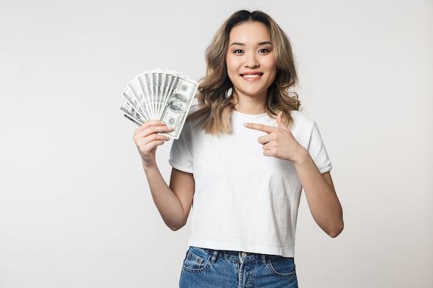 Portrait of a lovely young asian woman standing isolated over white wall, showing money banknotes