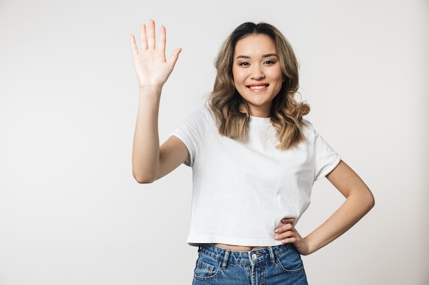 Portrait of a lovely young asian woman standing isolated over white wall, waving hand