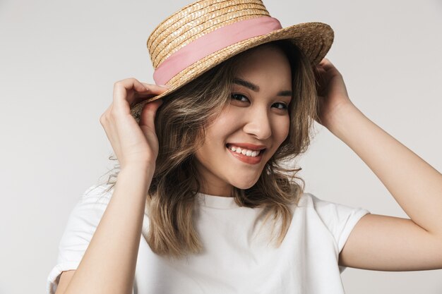 Portrait of a lovely young asian woman standing isolated over white wall, wearing summer hat, posing