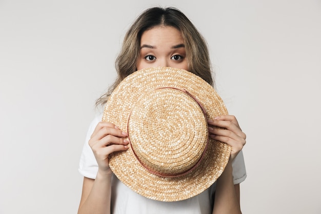 Foto ritratto di una giovane donna asiatica adorabile in piedi isolata su un muro bianco, con indosso un cappello estivo, in posa