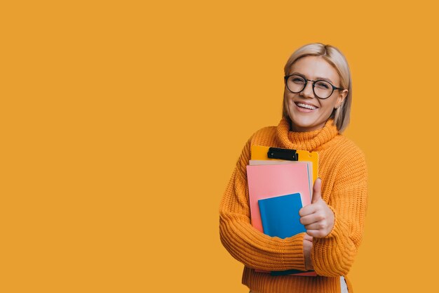 Portrait of a lovely young blonde female student looking at camera smiling while showing thumb up holding her book isolated on yellow studio wall.