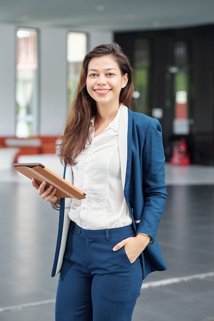 Portrait of lovely young business lady standing in office hall with digital tablet in hands