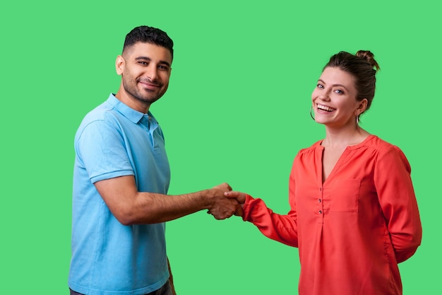 Portrait of lovely young couple in casual wear standing looking at camera with smile and shaking hands friendly meeting or acquaintance greeting isolated on green background indoor studio shot
