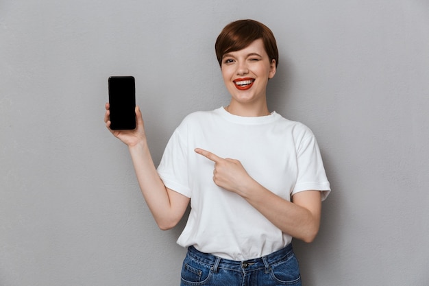 Портрет прекрасной молодой женщины, улыбающейся и указывающей пальцем на мобильный телефон, изолированной над серой стеной