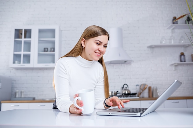 Portrait of lovely young woman studying at home on laptop. A girl with long blond hair in a sweater drinks tea. Modern kitchen interior and education concept. Work remotely from home