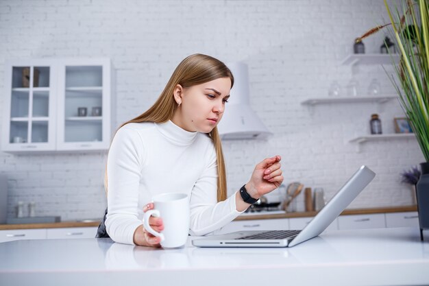 Portrait of lovely young woman studying at home on laptop. A girl with long blond hair in a sweater drinks tea. Modern kitchen interior and education concept. Work remotely from home