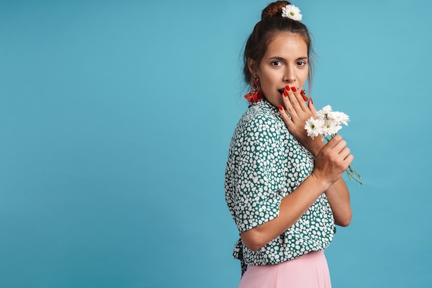 Portrait of a lovely young woman wearing shirt and bright earrings standing isolated over blue wall, holding chamomile bouquet