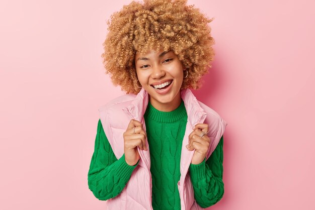 Portrait of lovely young woman with curly hair smiles toothily
wears green jumper and vest feels happy being in good mood isolated
over pink background smiling female model glad to meet friend