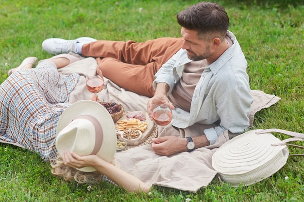 Portrait of loving adult couple enjoying picnic on green grass and drinking wine during romantic date outdoors