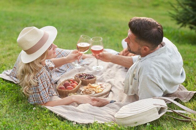 Ritratto di amorevole coppia adulta godendo picnic alla luce del sole e tintinnio di bicchieri di vino durante un appuntamento romantico all'aperto