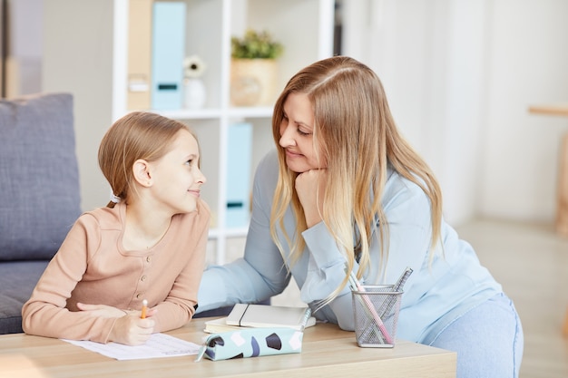 Portrait of loving adult mother looking at cute little girl doing homework while studying at home in cozy interior, copy space