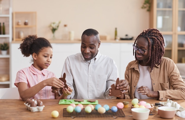 Портрет любящей афро-американской семьи, делающей шоколадных пасхальных кроликов, сидя за деревянным столом в уютном домашнем интерьере и наслаждаясь приготовлениями к празднику
