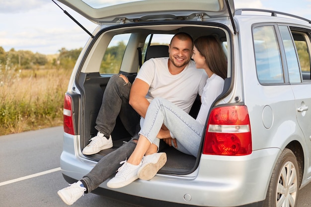 Portrait of loving couple sitting in car trunk wearing white shirts traveling together talking and smiling being fall in love enjoying spending time together