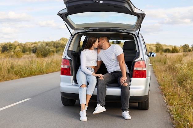 Portrait of loving couple sitting and kissing in their car in the trunk in nature on road man and woman wearing white shirts traveling together having romantic journey