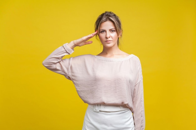 Portrait of loyal young woman with fair hair in casual beige blouse standing making salute with hand on head accepting order from boss indoor studio shot isolated on yellow background