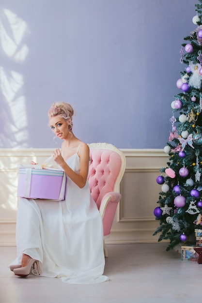 Portrait of a luxurious blonde in a white dress sitting on a chair by the christmas tree round gift box in hand .christmas.winter