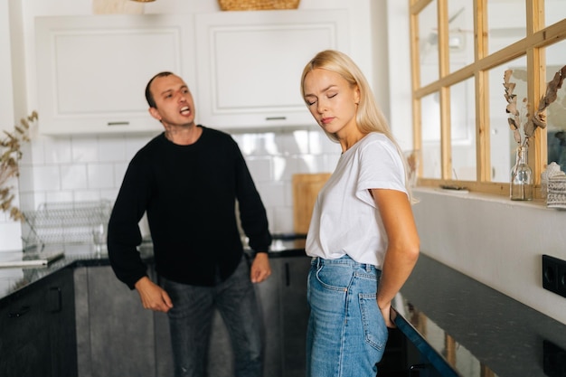 Portrait of mad emotional boyfriend scolding raising voice screaming at ignoring young wife wife standing in kitchen room