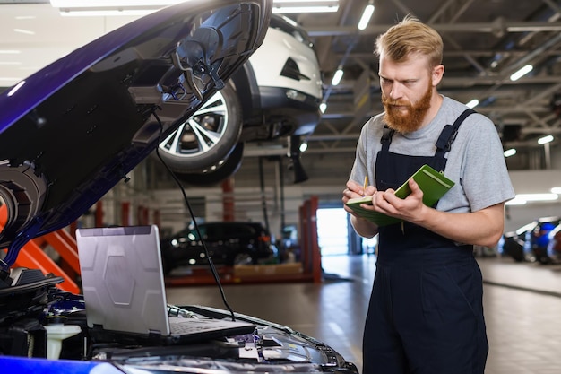Portrait of a male automotive electrician standing next to a diagnostic laptop and writing errors in a notebook Repair and maintenance of cars at a service station