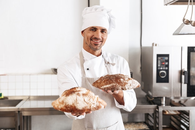 Portrait of male baker in white uniform standing at bakery, and holding bread