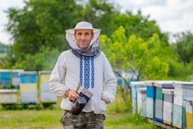 Portrait of male beekeeper with hives at background. Protective clothes on. Apiary.
