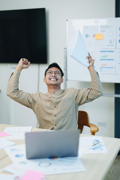 Portrait of a male business owner showing a happy smiling face as he has successfully invested his business using computers and financial budget documents at work