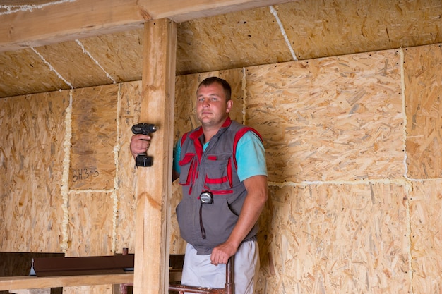 Ritratto di maschio operaio edile costruttore azienda trapano cordless all'interno di casa incompiuta con pannelli di particelle di compensato a vista