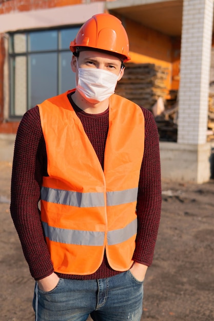 Portrait of male construction worker in medical mask and overalls on background of house under construction