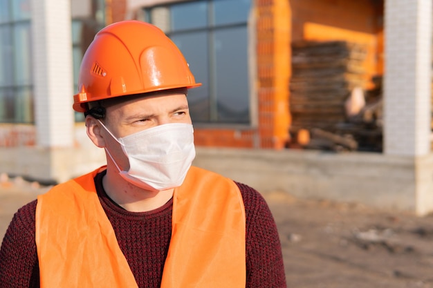 Ritratto di lavoratore edile maschile in maschera medica e tuta sullo sfondo della casa in costruzione