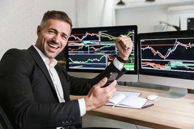 Portrait of male director 30s wearing suit using mobile phone while working with digital graphics on computer in office