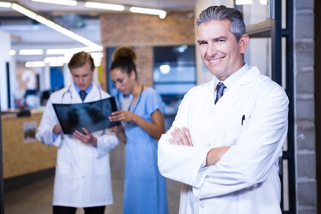 portrait of male doctor standing with arms crossed in hospital and colleagues standing behind and discussing