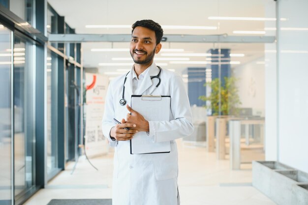 Portrait of male indian doctor wearing white coat having open door on clinic corridor as background