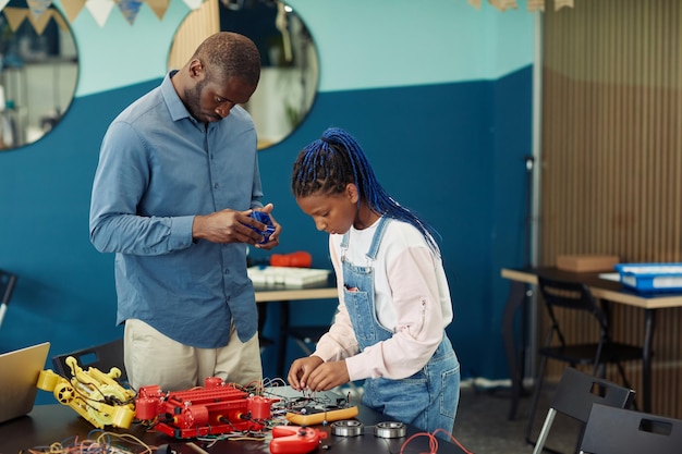 Portrait of male teacher helping young african american girl building robot during engineering class