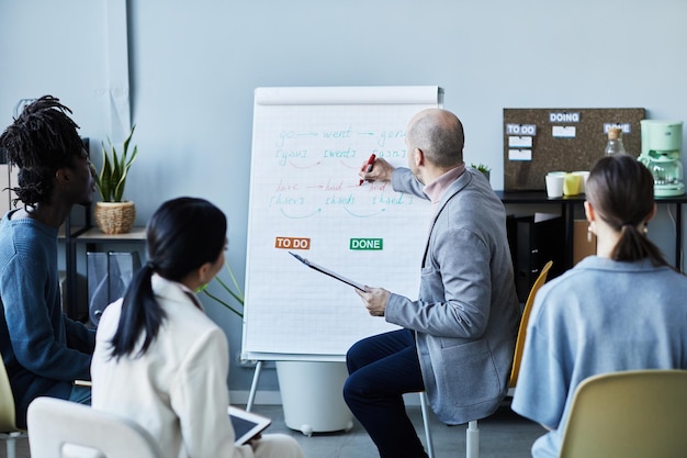 Photo portrait of male teacher writing on whiteboard while giving english lesson to group of people sittin