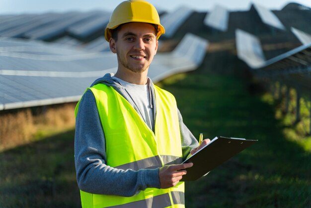 Portrait of male technologist in uniform with protective helmet\
adult man holding clip board in his hands standing on field with\
solar panels