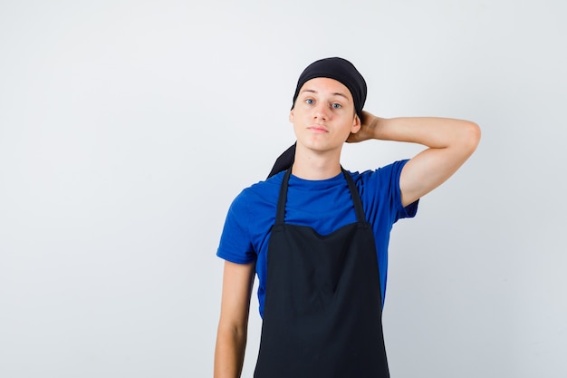 Portrait of male teen cook keeping hand behind head in t-shirt, apron and looking preoccupied front view