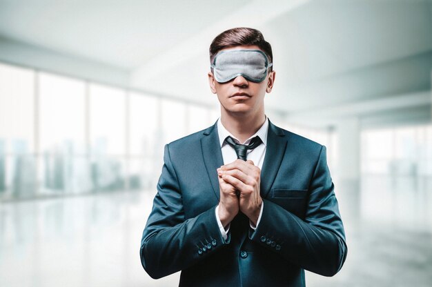 Portrait of a man in a business suit. He is praying while standing in the office. He has a sleep mask on his eyes. Blind business concept. 