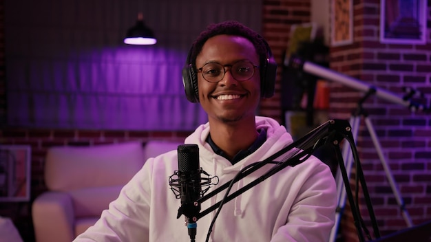 Portrait of man content creator smiling confident at camera\
while answering questions from listeners in home recording studio.\
african american online radio host reading fan mail from laptop\
computer.